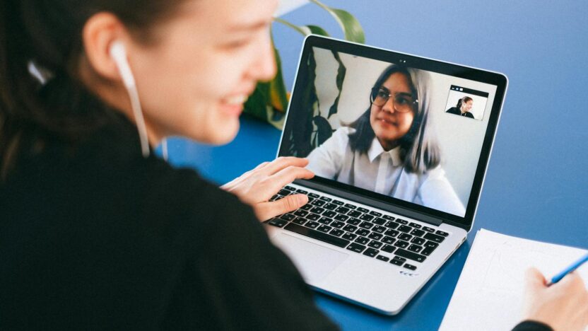 Woman having a video conference on a laptop, smiling and taking notes.